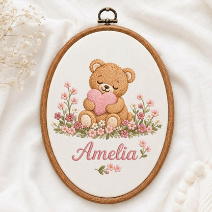 May include: An oval wooden embroidery hoop containing a fabric panel with a stitched design of a brown teddy bear holding a pink heart, surrounded by pink and white flowers. The name Amelia is embroidered in pink script below the bear.