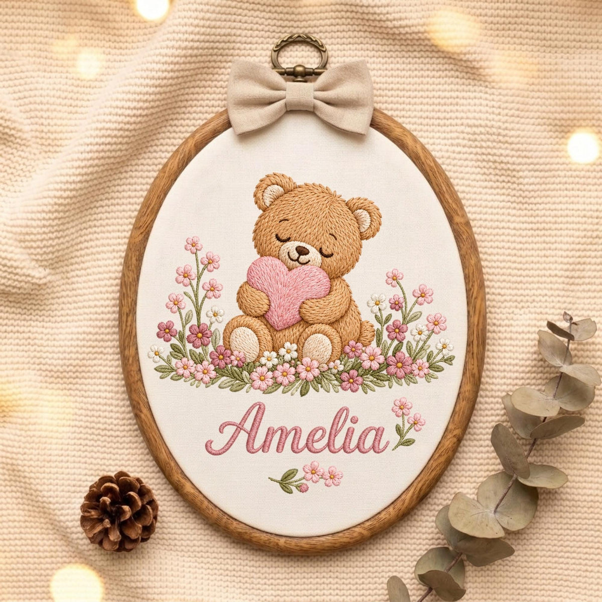 May include: An oval embroidery hoop featuring a brown teddy bear holding a pink heart, surrounded by pink and white flowers. The name Amelia is embroidered in pink script below the bear. A beige fabric bow is attached to the top metal hanger.