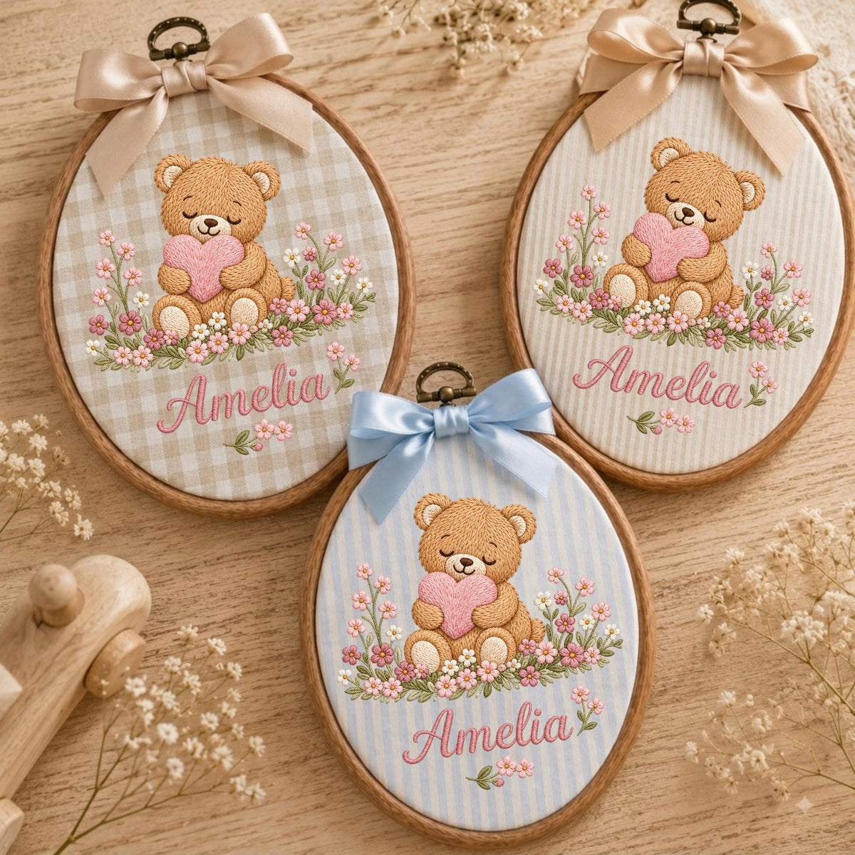 May include: Three oval wooden embroidery hoops featuring fabric backgrounds with a brown teddy bear holding a pink heart surrounded by pink flowers. Each hoop has a satin ribbon bow at the top and the name Amelia embroidered in pink script.