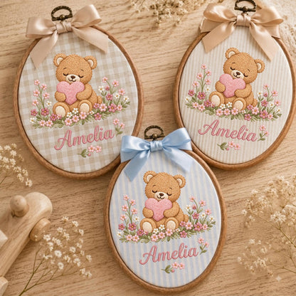 May include: Three oval wooden embroidery hoops featuring fabric backgrounds with a brown teddy bear holding a pink heart surrounded by pink flowers. Each hoop has a satin ribbon bow at the top and the name Amelia embroidered in pink script.