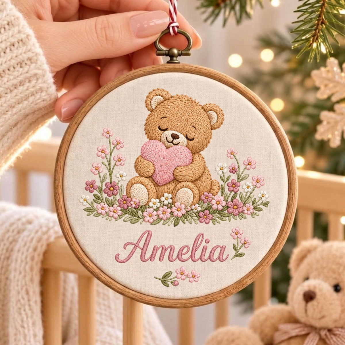 May include: A circular wooden embroidery hoop containing a cream-colored fabric with a stitched design of a brown teddy bear holding a pink heart, surrounded by pink and white flowers. The name Amelia is embroidered in pink script below the bear.