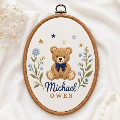 May include: An oval wooden embroidery hoop containing white fabric with a stitched brown teddy bear wearing a dark blue bow tie. The design includes blue floral stems, small gold and blue stars, and the embroidered names Michael and OWEN.