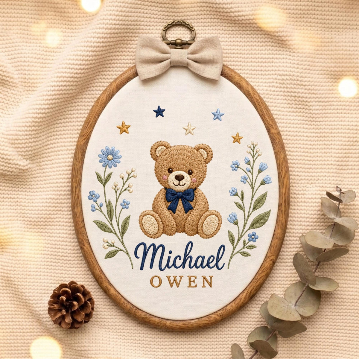 May include: An oval embroidery hoop featuring a brown teddy bear with a dark blue bow tie, flanked by blue floral sprigs and small stars. The name Michael OWEN is embroidered in dark blue and gold text. A beige fabric bow sits at the top.