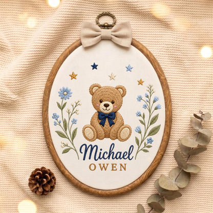 May include: An oval embroidery hoop featuring a brown teddy bear with a dark blue bow tie, flanked by blue floral sprigs and small stars. The name Michael OWEN is embroidered in dark blue and gold text. A beige fabric bow sits at the top.