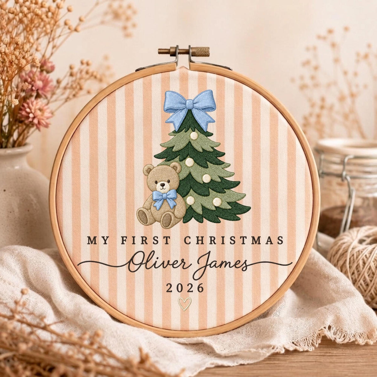 May include: A round wooden embroidery hoop featuring a beige and white striped fabric background. The design includes an embroidered green Christmas tree with a light blue bow, a brown teddy bear with a blue bow, and the text MY FIRST CHRISTMAS Oliver James 2026 with a small heart icon.