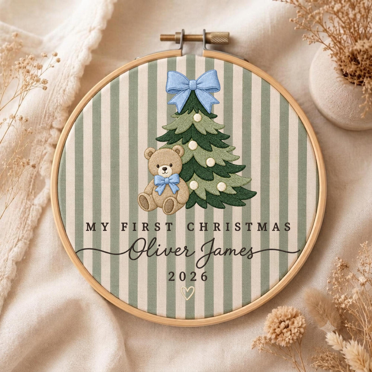 May include: A round wooden embroidery hoop containing fabric with vertical sage green and white stripes. The design features an embroidered green Christmas tree with a light blue bow, a brown teddy bear with a blue bow, and the text MY FIRST CHRISTMAS, Oliver James, 2026, and a small heart.