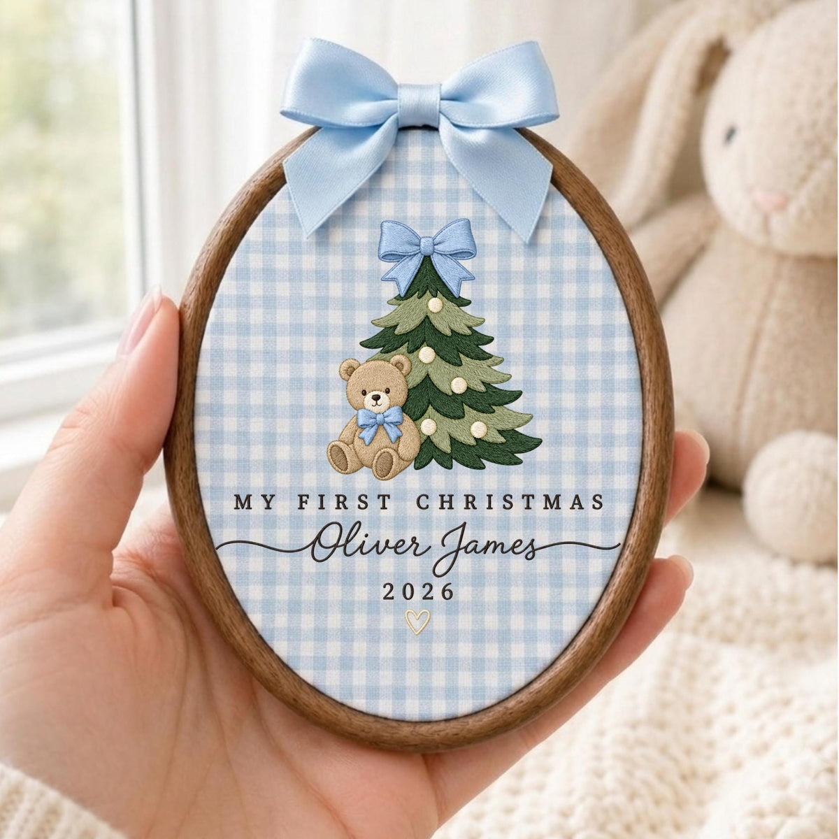 May include: An oval wooden embroidery hoop containing fabric with a light blue gingham pattern. It features an embroidered green Christmas tree, a teddy bear with a blue bow, and the text MY FIRST CHRISTMAS Oliver James 2026 with a heart.