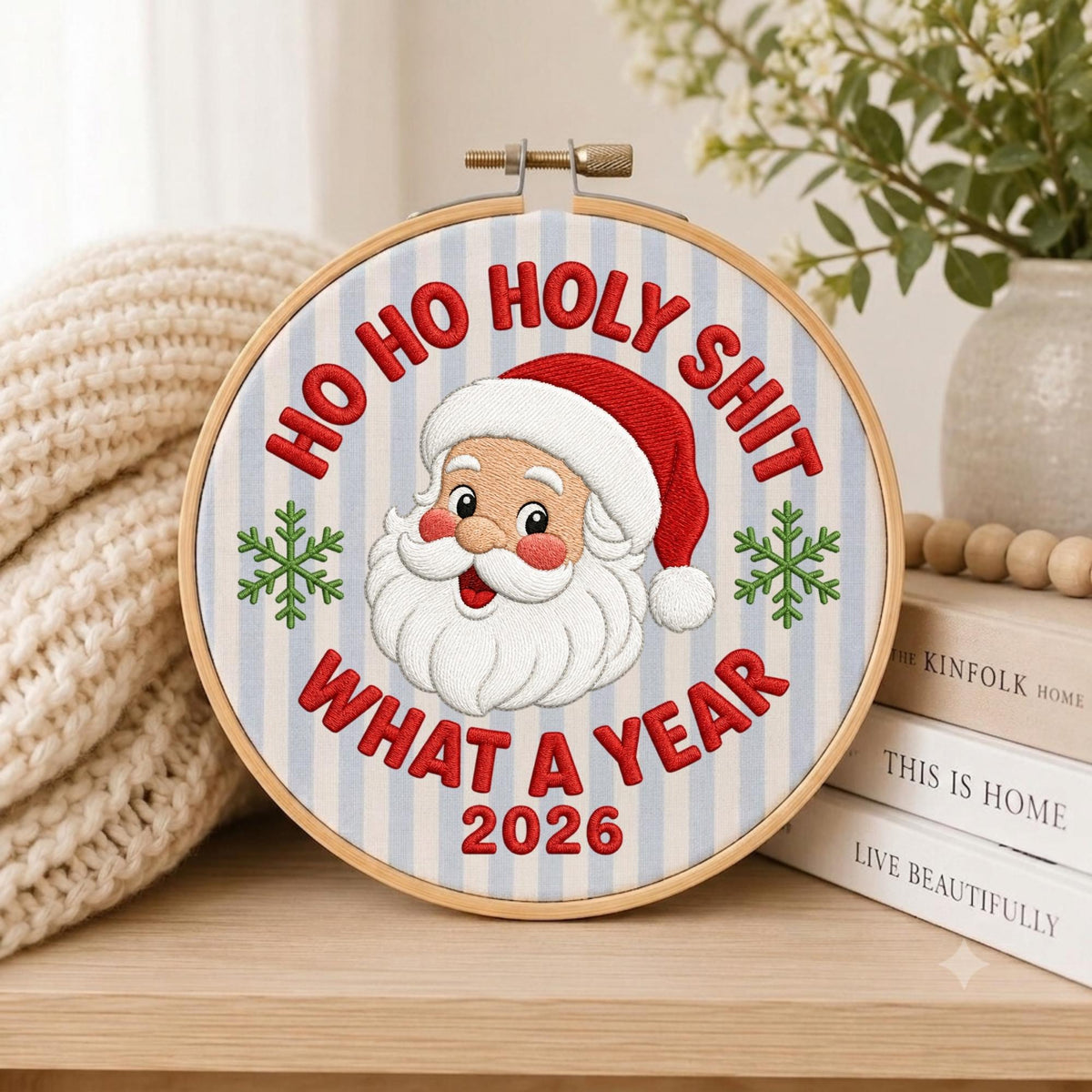 May include: A circular wooden embroidery hoop containing fabric with light blue vertical stripes. The embroidery features a smiling Santa Claus in a red hat, flanked by two green snowflakes, with the red text HO HO HOLY SHIT WHAT A YEAR 2026.
