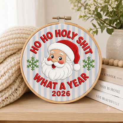 May include: A circular wooden embroidery hoop containing fabric with light blue vertical stripes. The embroidery features a smiling Santa Claus in a red hat, flanked by two green snowflakes, with the red text HO HO HOLY SHIT WHAT A YEAR 2026.