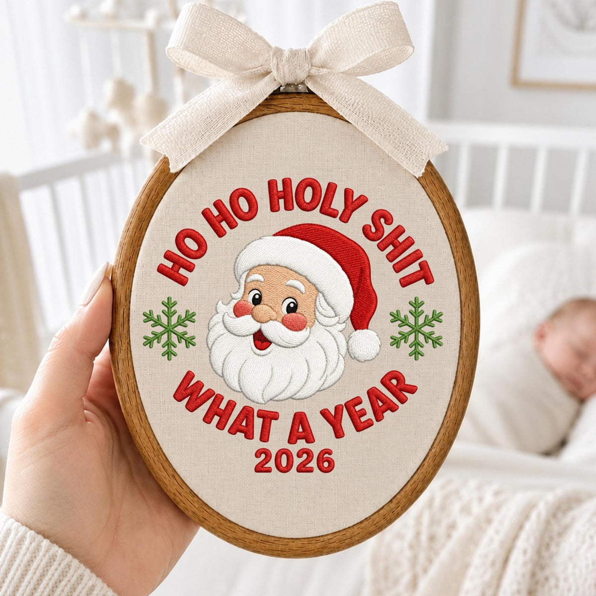 May include: An oval wooden embroidery hoop containing a cream-colored fabric with an embroidered Santa Claus face, green snowflakes, and red text that reads HO HO HOLY SHIT WHAT A YEAR 2026. A beige fabric bow is attached to the top of the hoop.