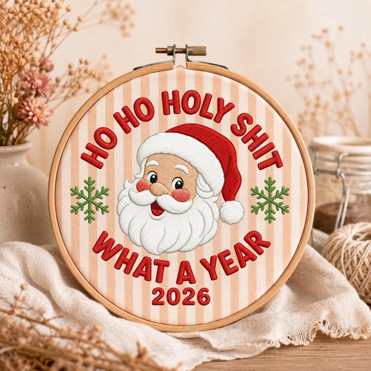 May include: A circular wooden embroidery hoop containing fabric with vertical beige stripes. The embroidery features a central Santa Claus face with a red hat, flanked by two green snowflakes. Red text reads: HO HO HOLY SHIT WHAT A YEAR 2026.