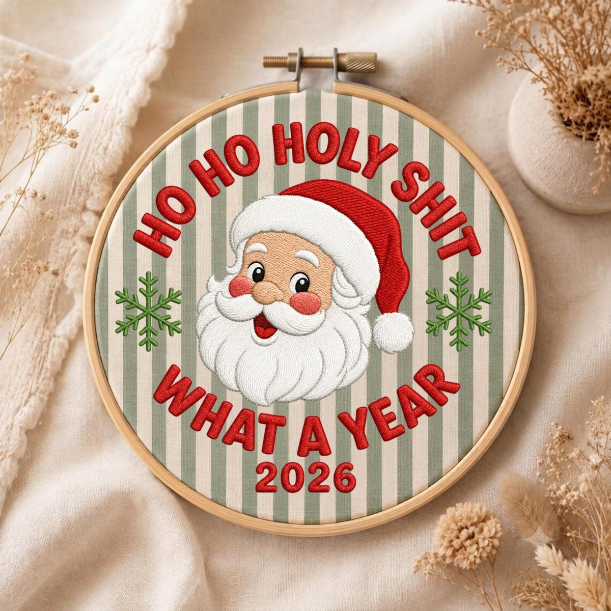 May include: A circular wooden embroidery hoop containing fabric with vertical light green and white stripes. The embroidery features a central Santa Claus face, two green snowflakes, and the red text HO HO HOLY SHIT WHAT A YEAR 2026.
