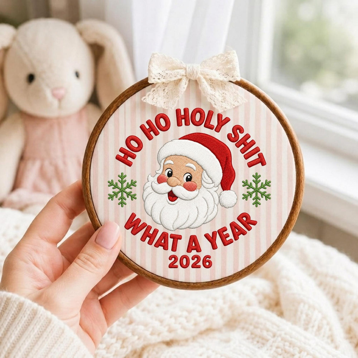 May include: A circular wooden embroidery hoop containing fabric with a striped pattern. The embroidery features a Santa Claus face, green snowflakes, and the red text "HO HO HOLY SHIT WHAT A YEAR 2026". A cream-colored lace bow is attached to the top.
