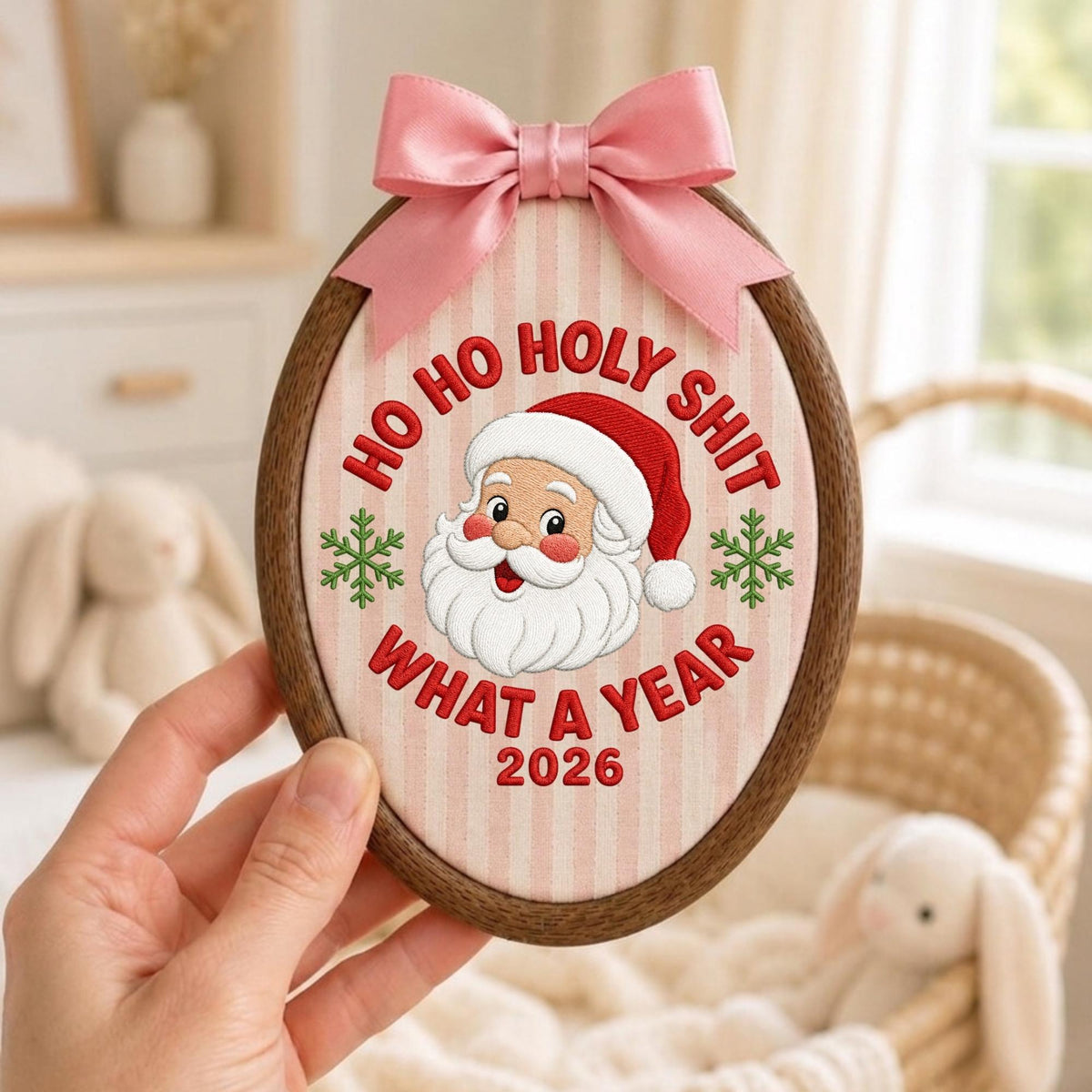 May include: An oval wooden embroidery hoop containing fabric with vertical light pink stripes. It features an embroidered Santa Claus face, green snowflakes, and the red text: HO HO HOLY SHIT WHAT A YEAR 2026. A pink satin ribbon bow is attached to the top.