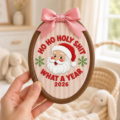 May include: An oval wooden embroidery hoop containing fabric with vertical light pink stripes. It features an embroidered Santa Claus face, green snowflakes, and the red text: HO HO HOLY SHIT WHAT A YEAR 2026. A pink satin ribbon bow is attached to the top.