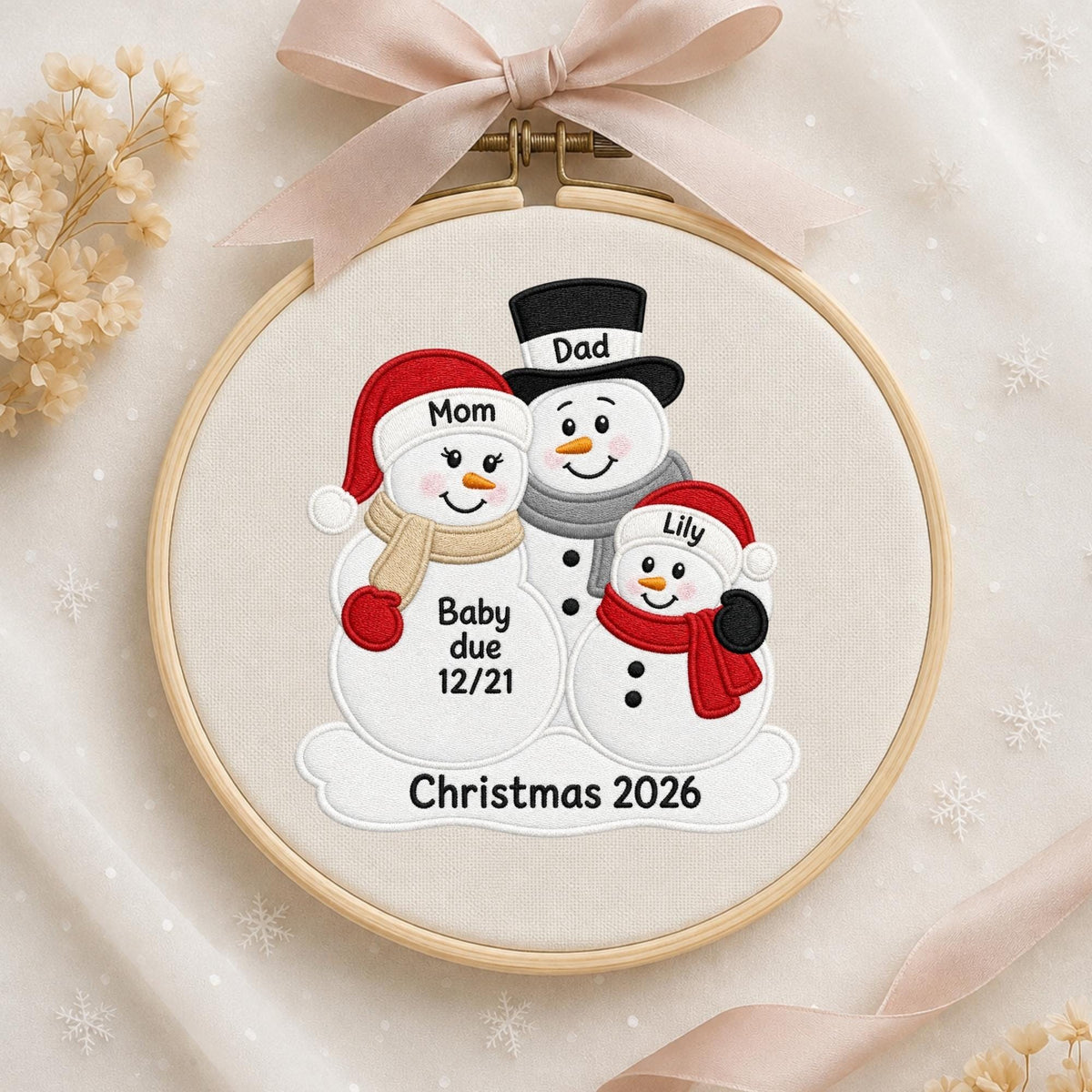 May include: A round wooden embroidery hoop containing a fabric panel with an embroidered snowman family. The design features three snowmen labeled Mom, Dad, and Lily, with the text Baby due 12/21 and Christmas 2026. A beige ribbon is tied at the top.
