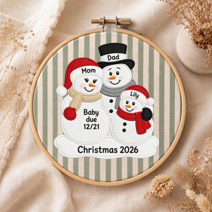 May include: A circular wooden embroidery hoop containing fabric with vertical sage green and white stripes. The fabric features an embroidered design of three snowmen with the text Mom, Dad, Lily, Baby due 12/21, and Christmas 2026.