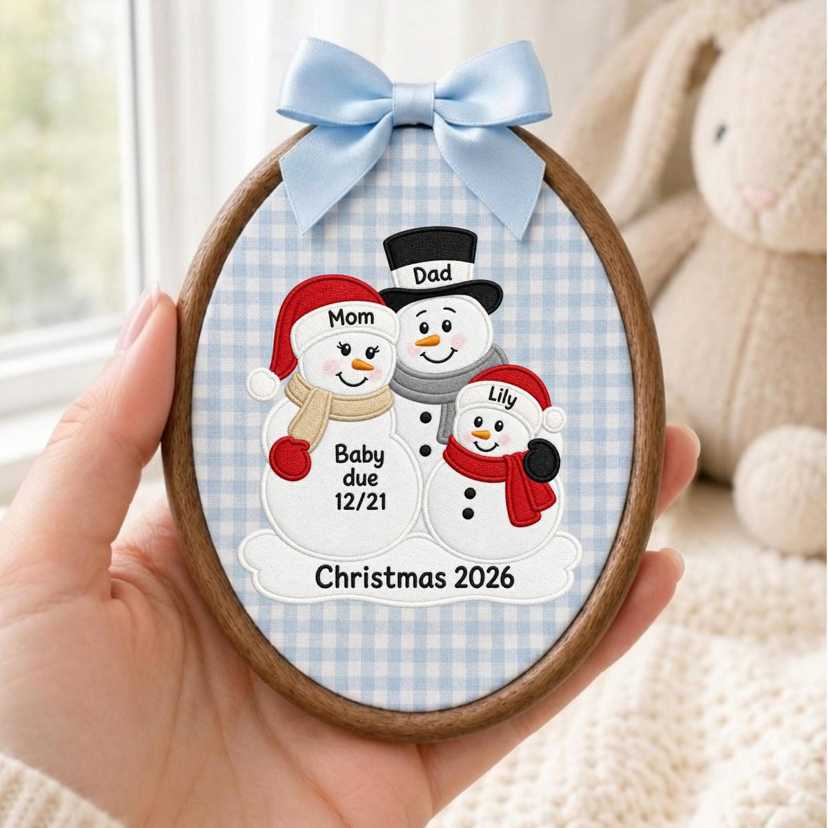 May include: An oval embroidery hoop ornament featuring a light blue and white gingham fabric background. The design shows three embroidered snowmen with the text Mom, Dad, Lily, Baby due 12/21, and Christmas 2026. A light blue satin bow is attached.