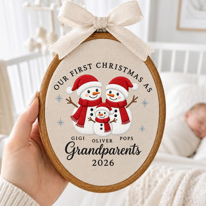 May include: An oval wooden embroidery hoop containing a fabric panel with a printed design of three snowmen wearing red hats and scarves. The text reads Our First Christmas As Grandparents, Gigi, Oliver, Pops, and 2026. A beige ribbon bow is tied at the top.