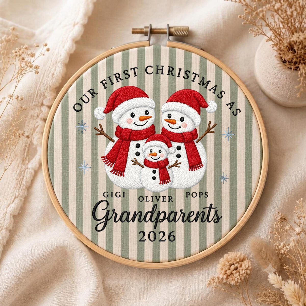 May include: A round wooden embroidery hoop containing fabric with a green and white striped pattern. The design features three snowmen wearing red hats and scarves, with the text "OUR FIRST CHRISTMAS AS GIGI OLIVER POPS Grandparents 2026".