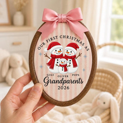 May include: An oval wooden embroidery hoop ornament featuring a pink and white striped fabric background. It displays three illustrated snowmen wearing red hats and scarves, with the text "OUR FIRST CHRISTMAS AS GIGI OLIVER POPS GRANDPARENTS 2026" and a pink satin bow at the top.