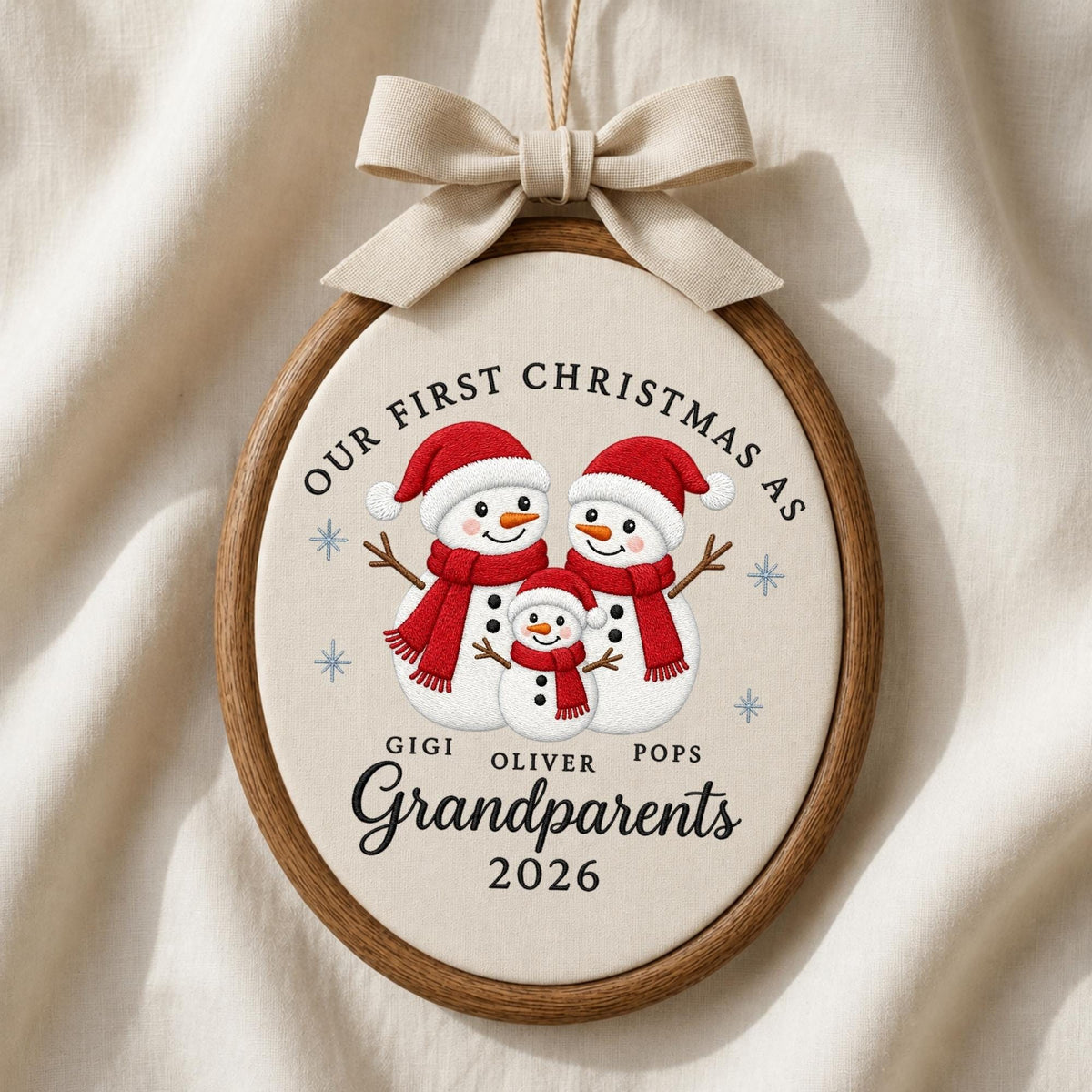 May include: An oval wooden embroidery hoop ornament featuring a cream fabric center with three embroidered snowmen wearing red hats and scarves. Text reads: Our First Christmas As Gigi Oliver Pops Grandparents 2026. A beige ribbon bow is attached.