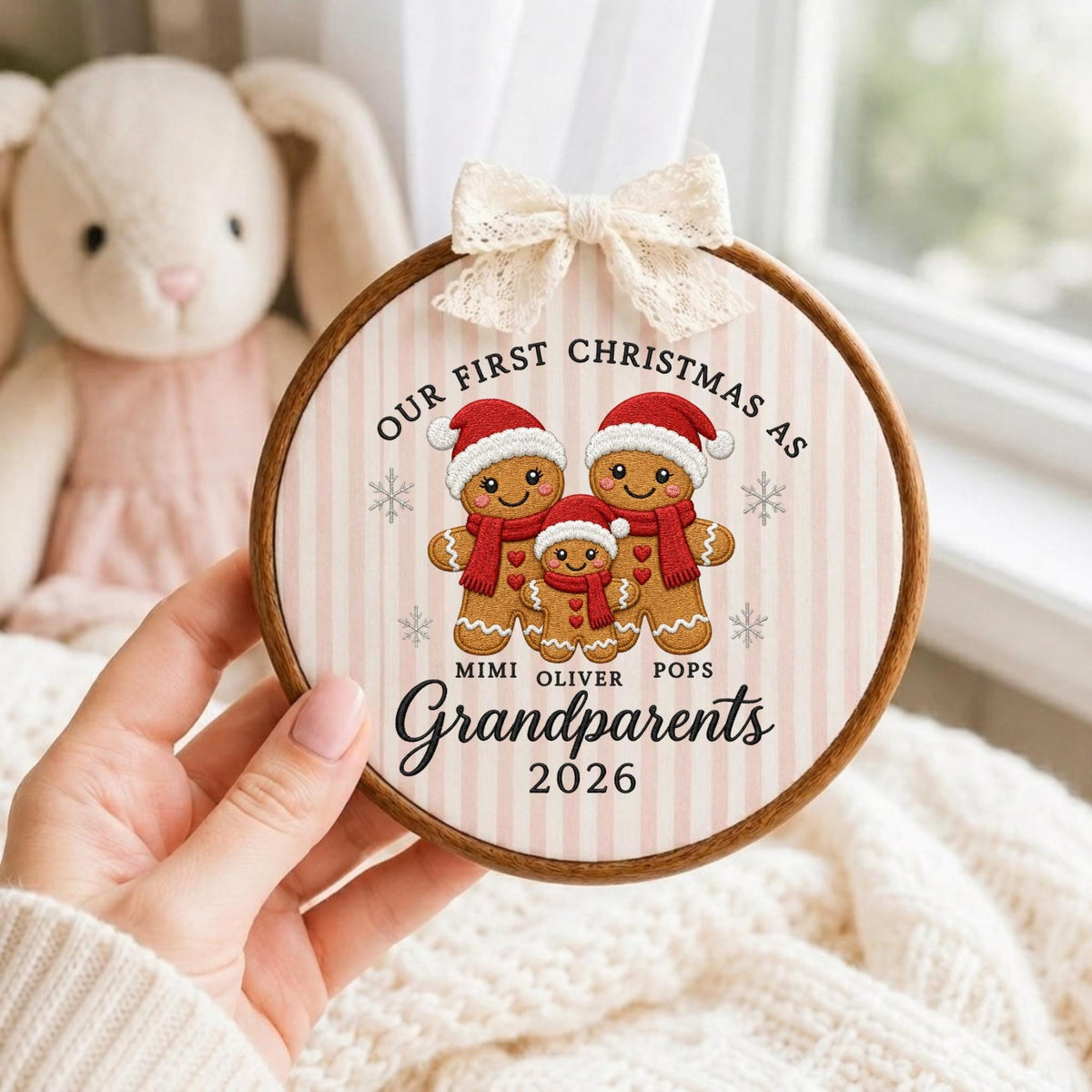 May include: A round wooden embroidery hoop ornament featuring a light pink and white striped fabric background. The design includes three gingerbread figures wearing red hats and scarves, with the text "OUR FIRST CHRISTMAS AS MIMI OLIVER POPS Grandparents 2026" and a cream-colored lace bow at the top.