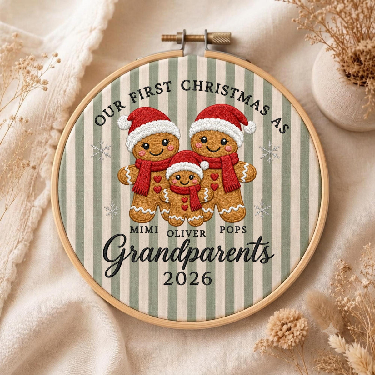 May include: A round wooden embroidery hoop containing fabric with a green and white vertical striped pattern. The design features three embroidered gingerbread people wearing red hats and scarves, with the text "OUR FIRST CHRISTMAS AS MIMI OLIVER POPS Grandparents 2026" printed in black.