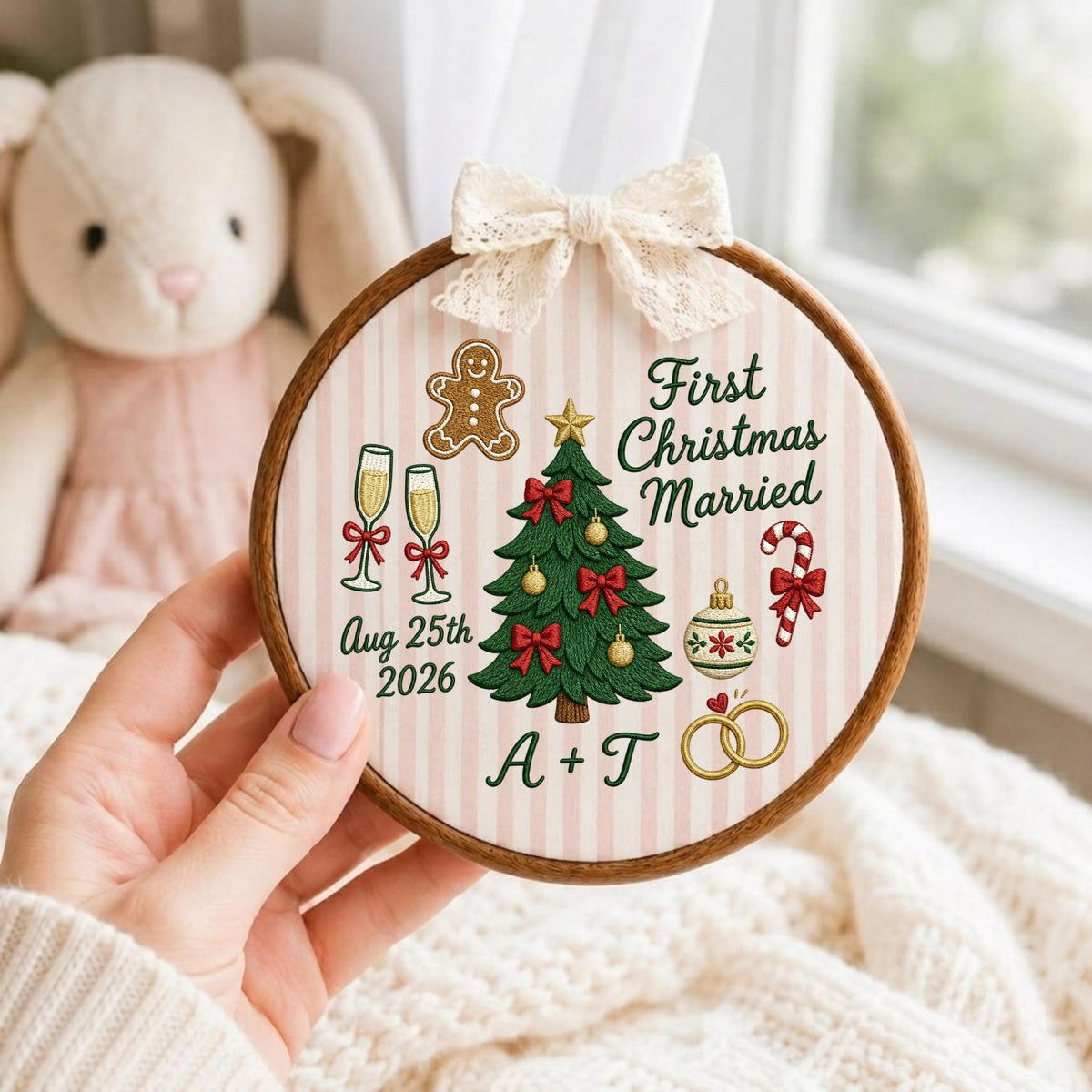 May include: A round wooden embroidery hoop featuring a light pink and white striped fabric background. The design includes a green Christmas tree, a gingerbread man, champagne flutes, a candy cane, and two gold rings. Text reads First Christmas Married, Aug 25th 2026, and A + T, topped with a cream lace bow.