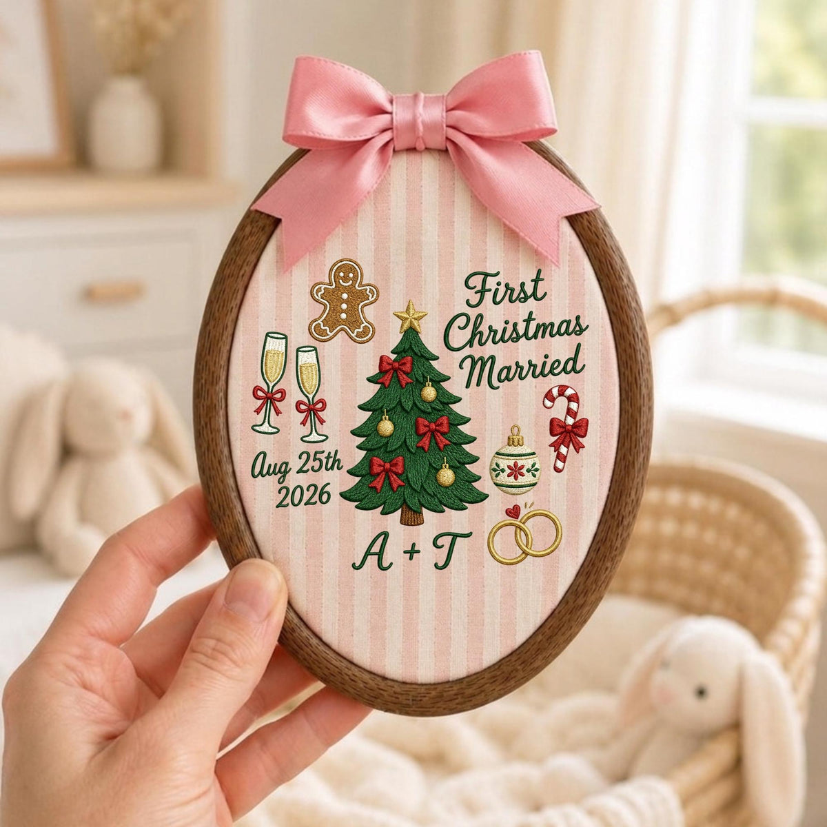 May include: An oval wooden embroidery hoop containing fabric with pink vertical stripes. The embroidery features a Christmas tree, gingerbread man, champagne flutes, candy cane, and rings with the text First Christmas Married, Aug 25th 2026, and A + T. A pink satin bow is attached to the top.