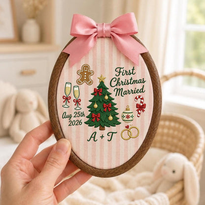 May include: An oval wooden embroidery hoop containing fabric with pink vertical stripes. The embroidery features a Christmas tree, gingerbread man, champagne flutes, candy cane, and rings with the text First Christmas Married, Aug 25th 2026, and A + T. A pink satin bow is attached to the top.