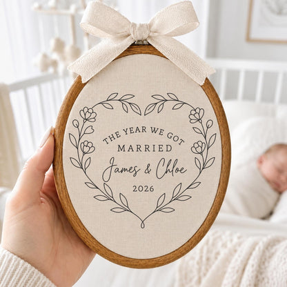 May include: An oval wooden embroidery hoop containing off-white fabric with black printed text that reads THE YEAR WE GOT MARRIED James &amp; Chloe 2026 inside a floral heart wreath. A beige fabric bow is attached to the top of the wooden frame.