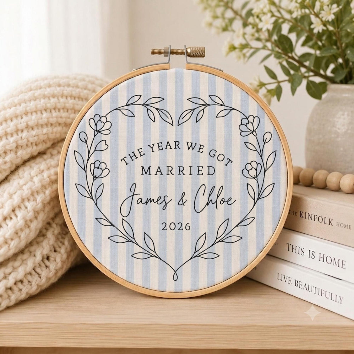 May include: A round wooden embroidery hoop framing light blue and white striped fabric. The fabric features a dark blue embroidered heart-shaped floral wreath surrounding the text: THE YEAR WE GOT MARRIED, James &amp; Chloe, and 2026.
