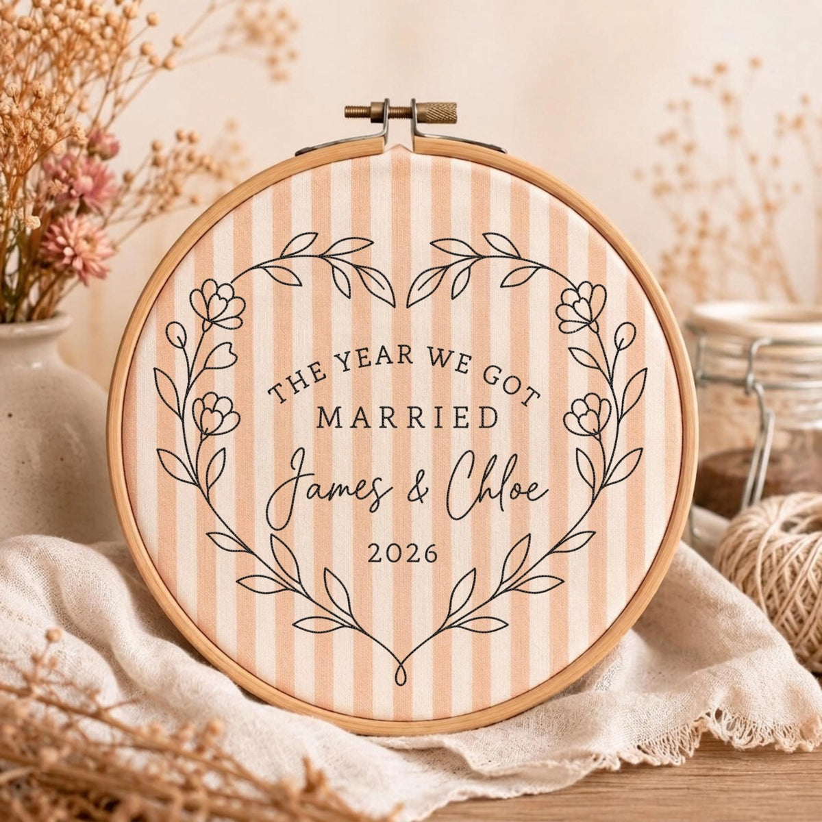 May include: A circular wooden embroidery hoop framing light peach and white striped fabric. The fabric features a black printed floral wreath surrounding the text THE YEAR WE GOT MARRIED, the names James &amp; Chloe, and the year 2026 in black ink.