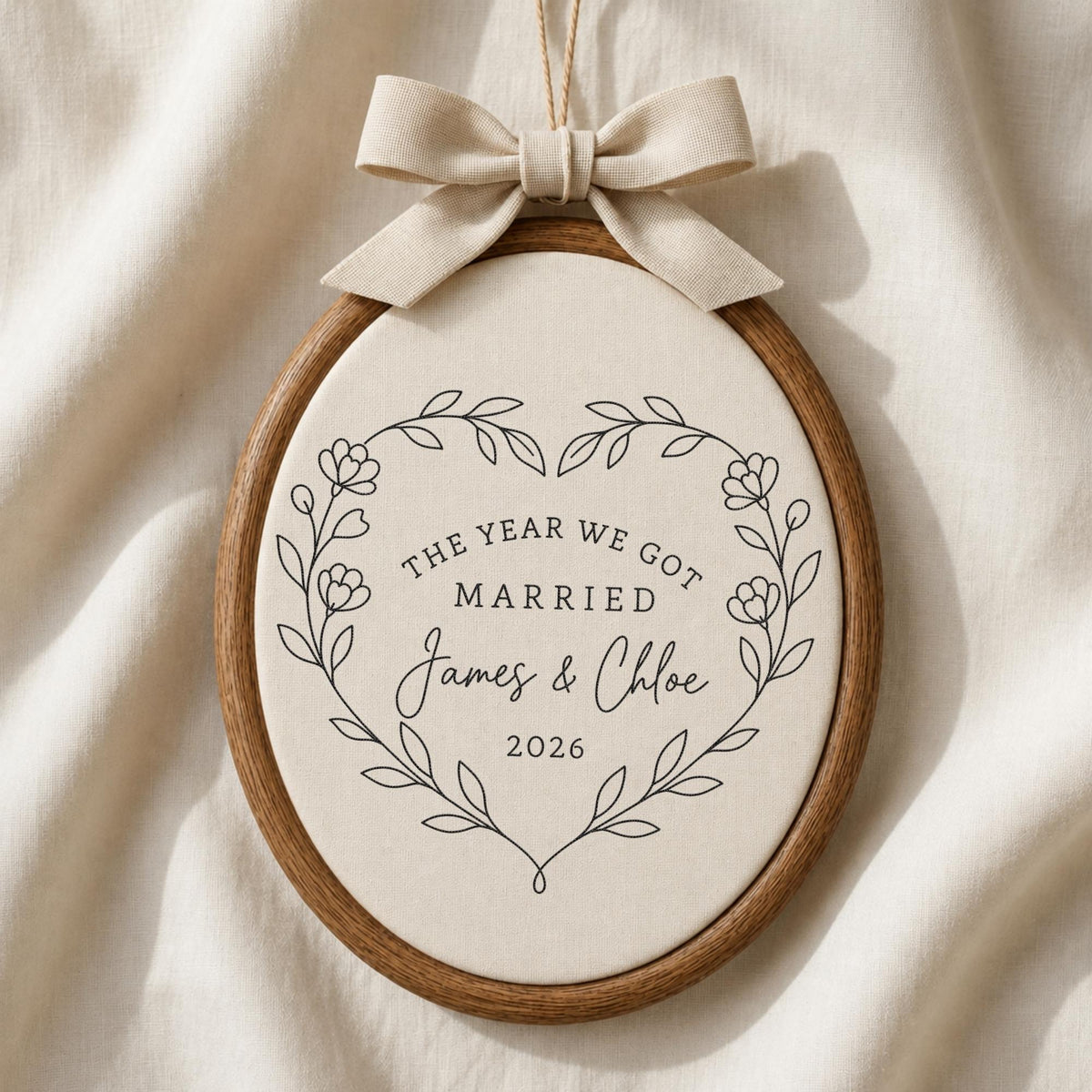May include: An oval wooden embroidery hoop frame containing off-white fabric with black printed text that reads THE YEAR WE GOT MARRIED James &amp; Chloe 2026 inside a floral heart wreath. A beige fabric bow is attached to the top of the frame.