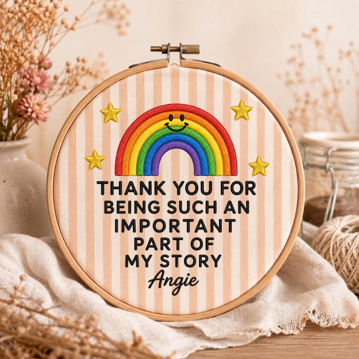 May include: A round wooden embroidery hoop containing fabric with vertical light pink and white stripes. The fabric features a colorful embroidered rainbow with a smiling face, four yellow stars, and the text: THANK YOU FOR BEING SUCH AN IMPORTANT PART OF MY STORY Angie.
