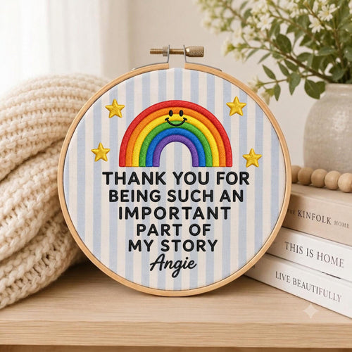 May include: A circular embroidery hoop featuring a light blue and white striped fabric background. An embroidered rainbow with a smiling face sits above the black text: THANK YOU FOR BEING SUCH AN IMPORTANT PART OF MY STORY Angie, with yellow stars.