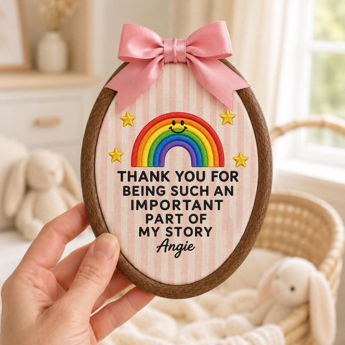 May include: An oval wooden embroidery hoop containing fabric with a light pink and white vertical stripe pattern. The fabric features an embroidered rainbow with a smiley face, four yellow stars, and the text: THANK YOU FOR BEING SUCH AN IMPORTANT PART OF MY STORY Angie. A pink satin ribbon bow is attached to the top.