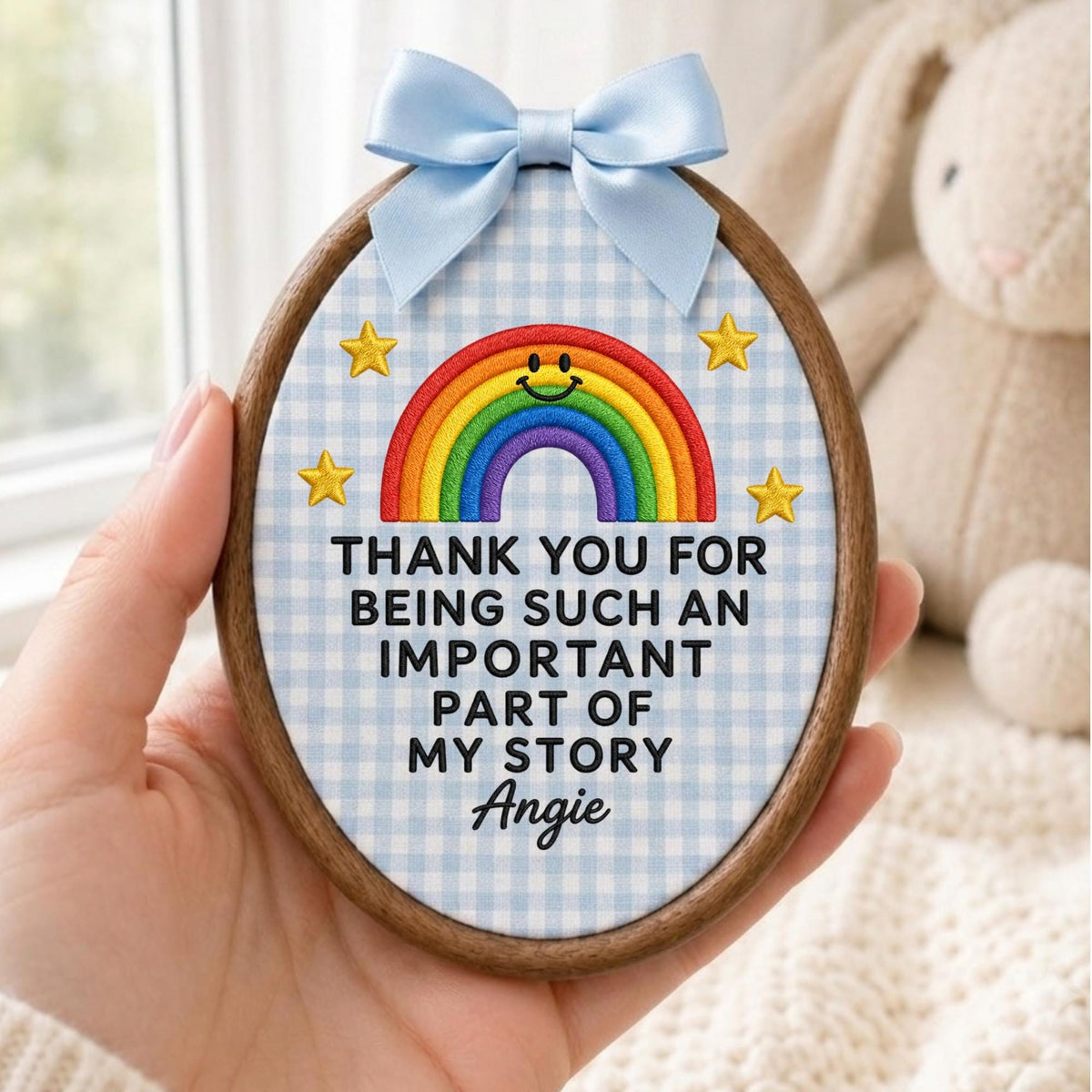 May include: An oval embroidery hoop frame containing fabric with a light blue gingham pattern. It features a colorful embroidered rainbow with a smiley face, four yellow stars, and the text: THANK YOU FOR BEING SUCH AN IMPORTANT PART OF MY STORY Angie. A light blue satin ribbon bow is attached to the top.