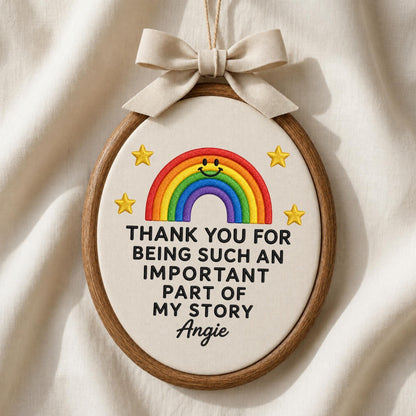 May include: An oval embroidery hoop containing off-white fabric with a stitched rainbow featuring a smiley face, four yellow stars, and the black text: THANK YOU FOR BEING SUCH AN IMPORTANT PART OF MY STORY Angie. A beige ribbon bow is at the top.