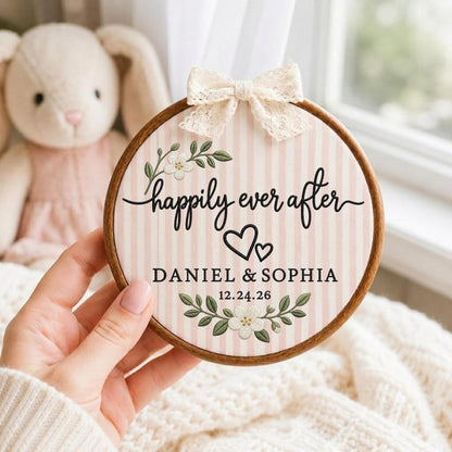 May include: A circular wooden embroidery hoop containing fabric with pink and white vertical stripes. The fabric features embroidered white flowers, green leaves, two black hearts, and the text "happily ever after DANIEL &amp; SOPHIA 12.24.26". A cream lace bow is attached to the top.