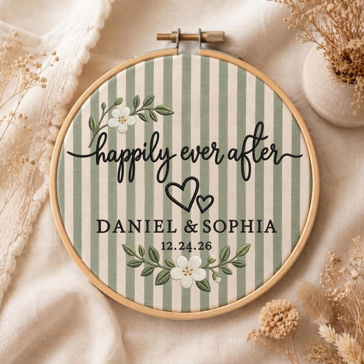 May include: A circular wooden embroidery hoop containing fabric with vertical sage green and cream stripes. The embroidery features the black text "happily ever after", "DANIEL &amp; SOPHIA", and "12.24.26", accented by two black hearts and white floral sprigs.