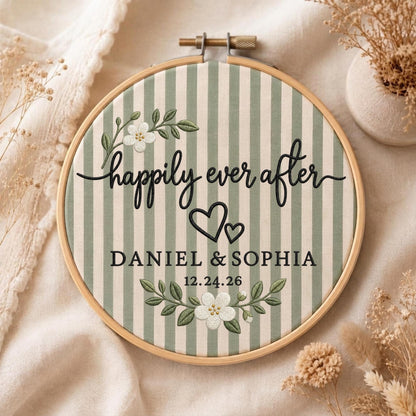 May include: A circular wooden embroidery hoop containing fabric with vertical sage green and cream stripes. The embroidery features the black text "happily ever after", "DANIEL &amp; SOPHIA", and "12.24.26", accented by two black hearts and white floral sprigs.