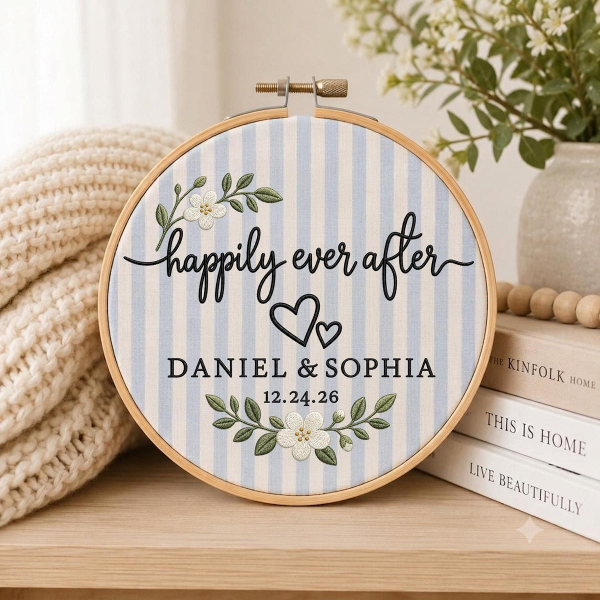 May include: A round wooden embroidery hoop containing fabric with light blue and white vertical stripes. The fabric features black embroidered text reading "happily ever after", "DANIEL &amp; SOPHIA", and "12.24.26", with two black hearts and white floral sprigs.