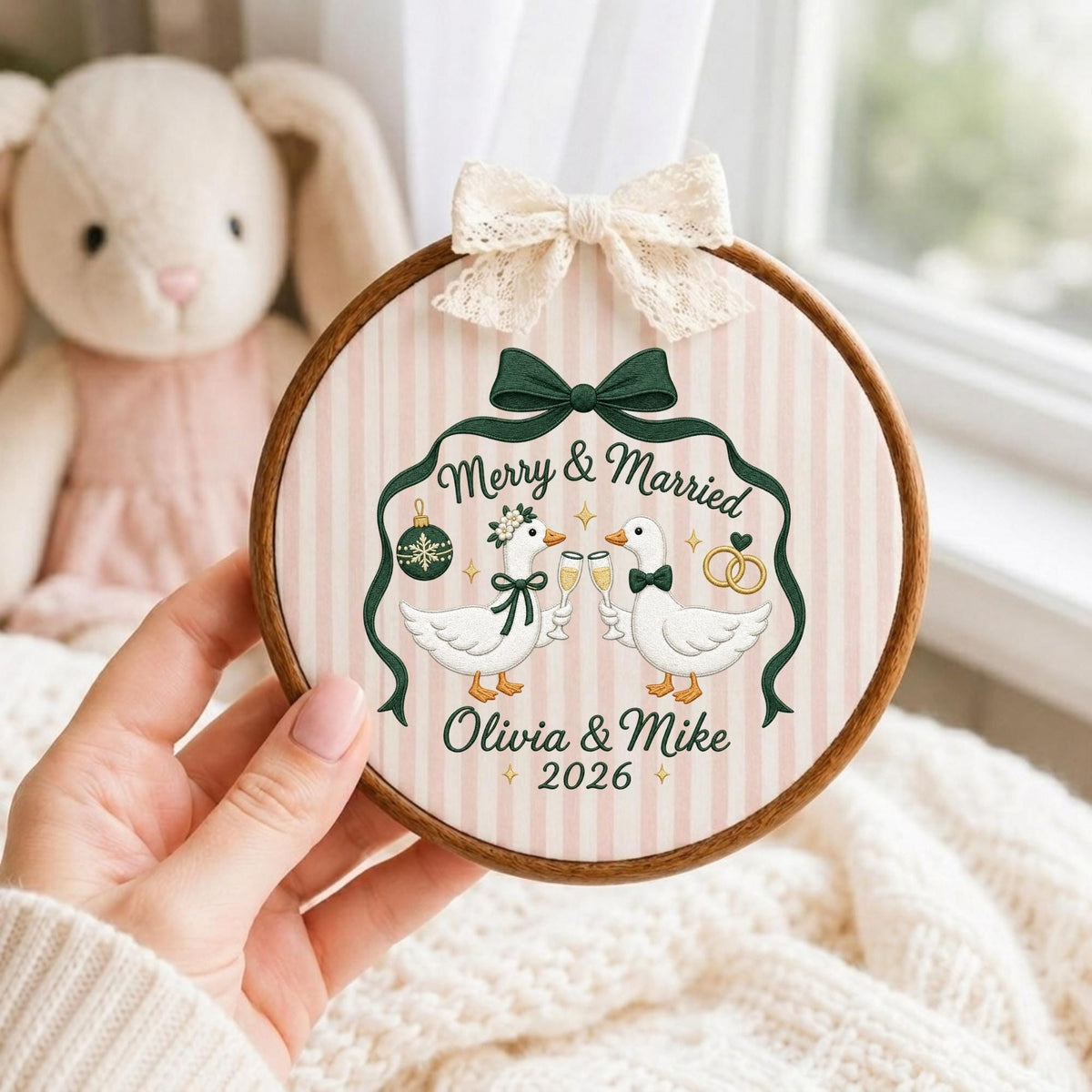 May include: A circular wooden embroidery hoop containing fabric with a pink and white striped pattern. The design features two white geese clinking glasses, a green bow, a Christmas ornament, and two gold rings. Text reads Merry &amp; Married, Olivia &amp; Mike, 2026. A cream lace bow is attached to the top.