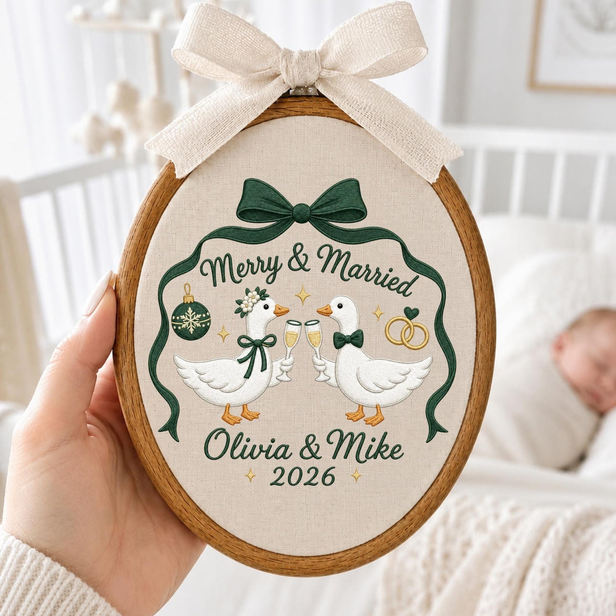 May include: An oval embroidery hoop featuring a beige fabric background with a dark green ribbon design. The embroidery depicts two white geese clinking champagne glasses, with the text Merry &amp; Married, Olivia &amp; Mike, and the year 2026.
