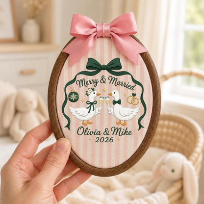 May include: An oval wooden embroidery hoop containing fabric with a pink and white striped pattern. The design features two white geese clinking champagne glasses, a green bow, a green ornament, gold rings, and the text Merry &amp; Married Olivia &amp; Mike 2026. A pink satin bow is attached to the top.