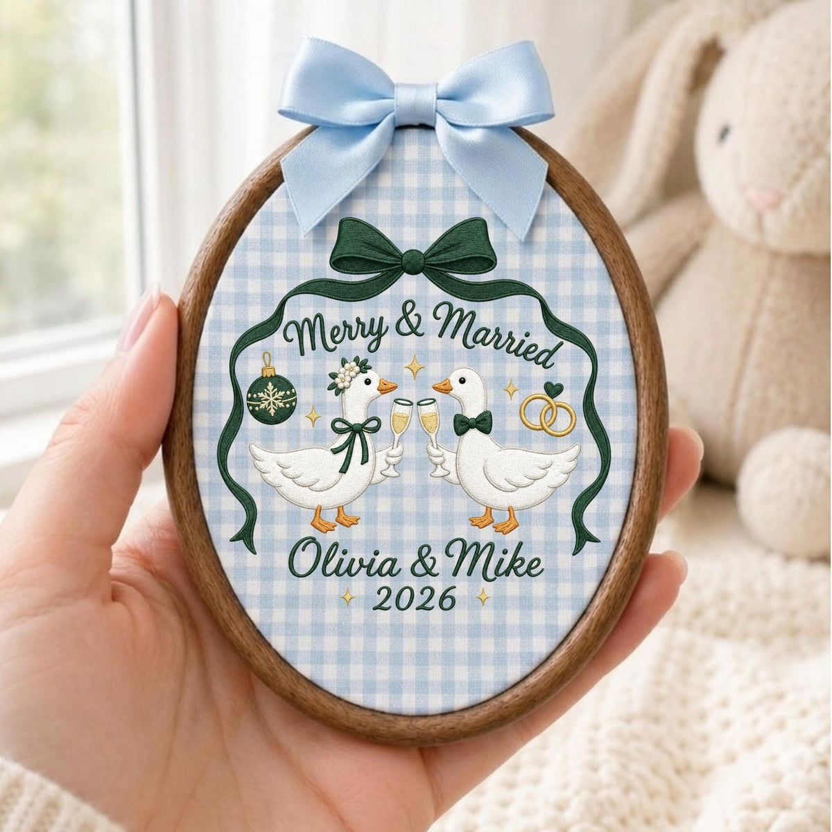 May include: An oval wooden embroidery hoop featuring light blue gingham fabric. The embroidery depicts two white geese holding champagne flutes, a green ornament, and gold rings. Text reads Merry &amp; Married, Olivia &amp; Mike, and 2026. A light blue satin bow is attached to the top.