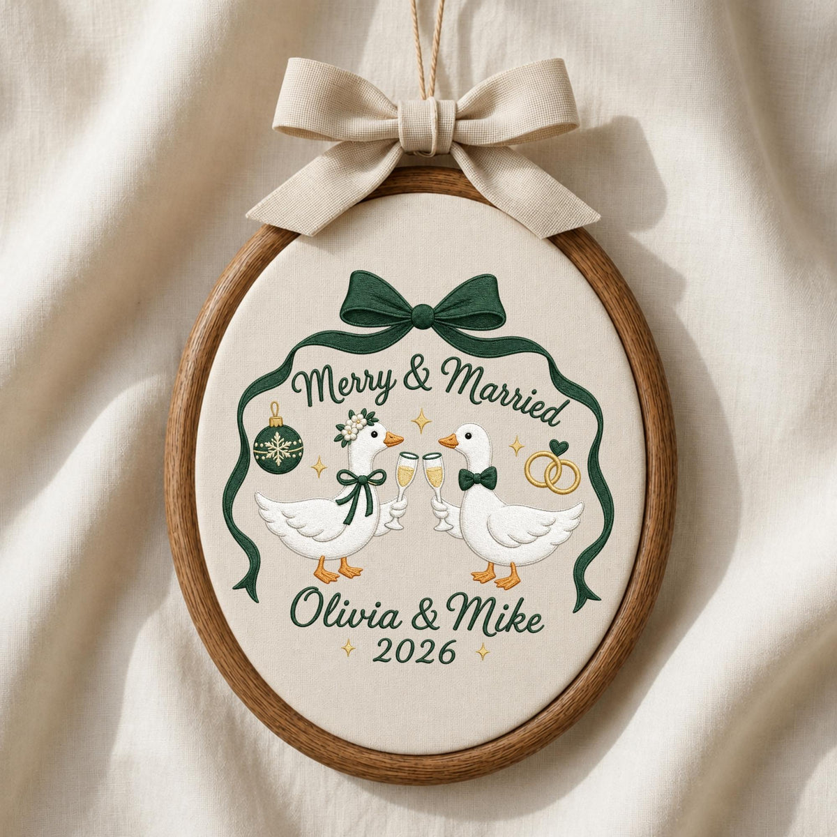 May include: An oval embroidery hoop containing fabric with a stitched design of two white geese clinking champagne glasses. The text reads Merry &amp; Married, Olivia &amp; Mike, and 2026. A dark green ribbon frames the geese, topped with a beige fabric bow.