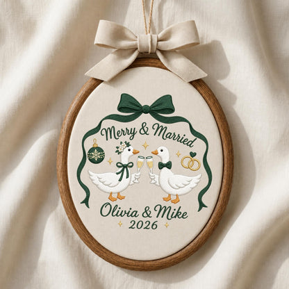 May include: An oval embroidery hoop containing fabric with a stitched design of two white geese clinking champagne glasses. The text reads Merry &amp; Married, Olivia &amp; Mike, and 2026. A dark green ribbon frames the geese, topped with a beige fabric bow.